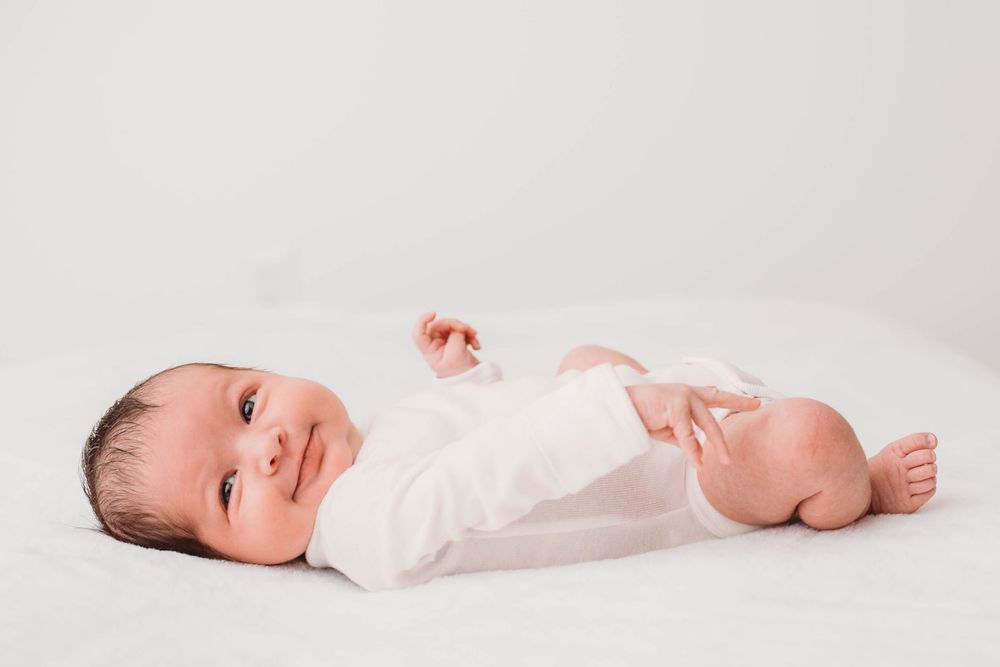 smiling baby wearing a white onesie laying in a white bed by beryl photography