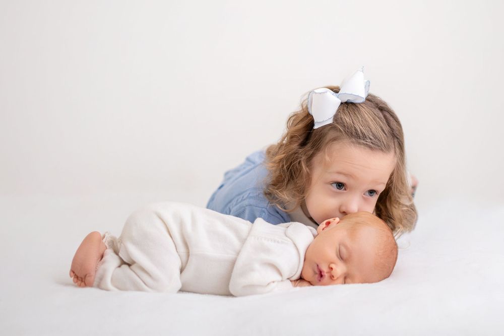 little girl in a blue dress with a white bow on her hair kissing a sleeping baby by beryl photography