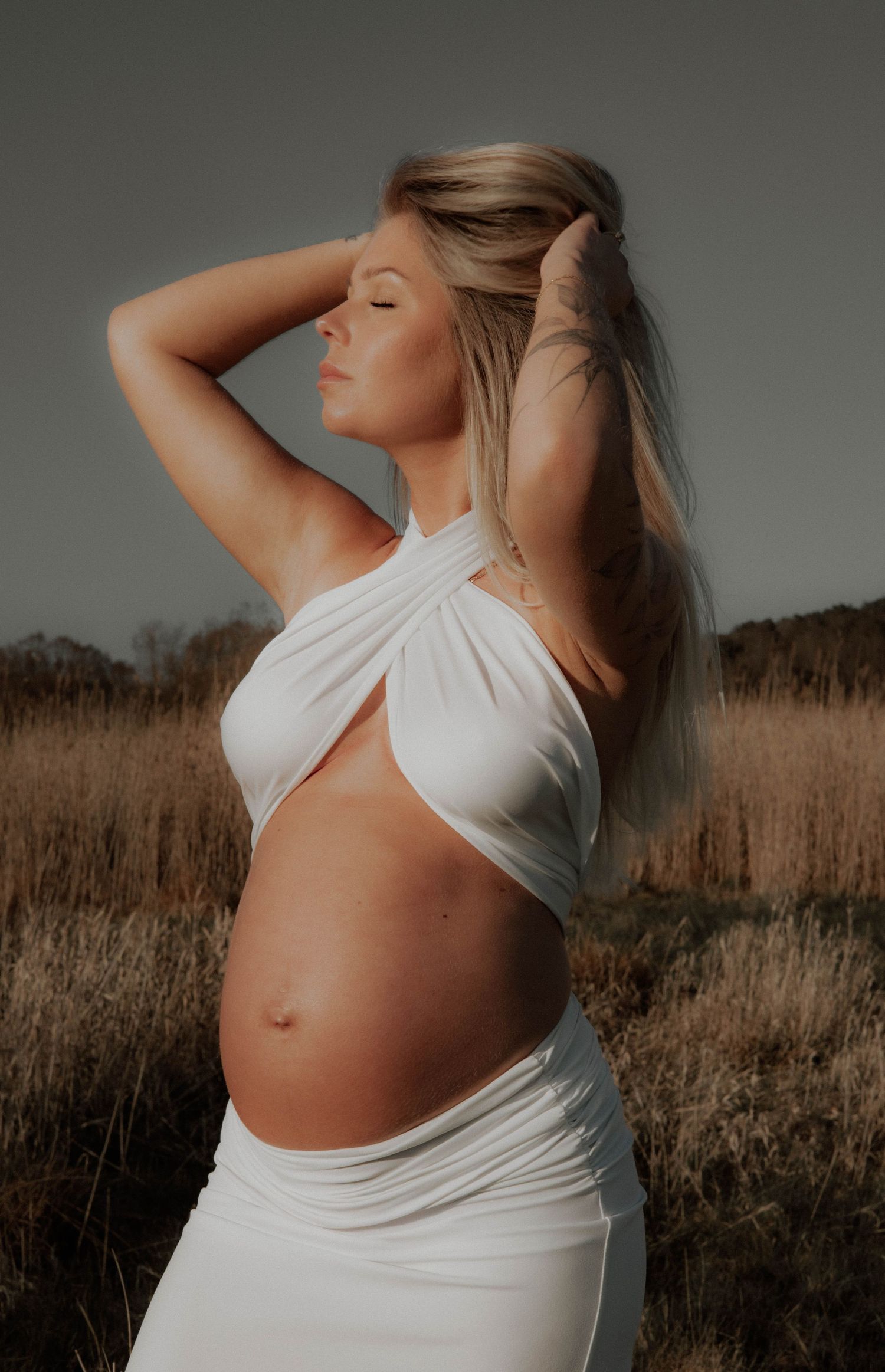 gravid kvinna står på en äng iklädd en vit tyllklänning. hon håller om sin gravidmage.