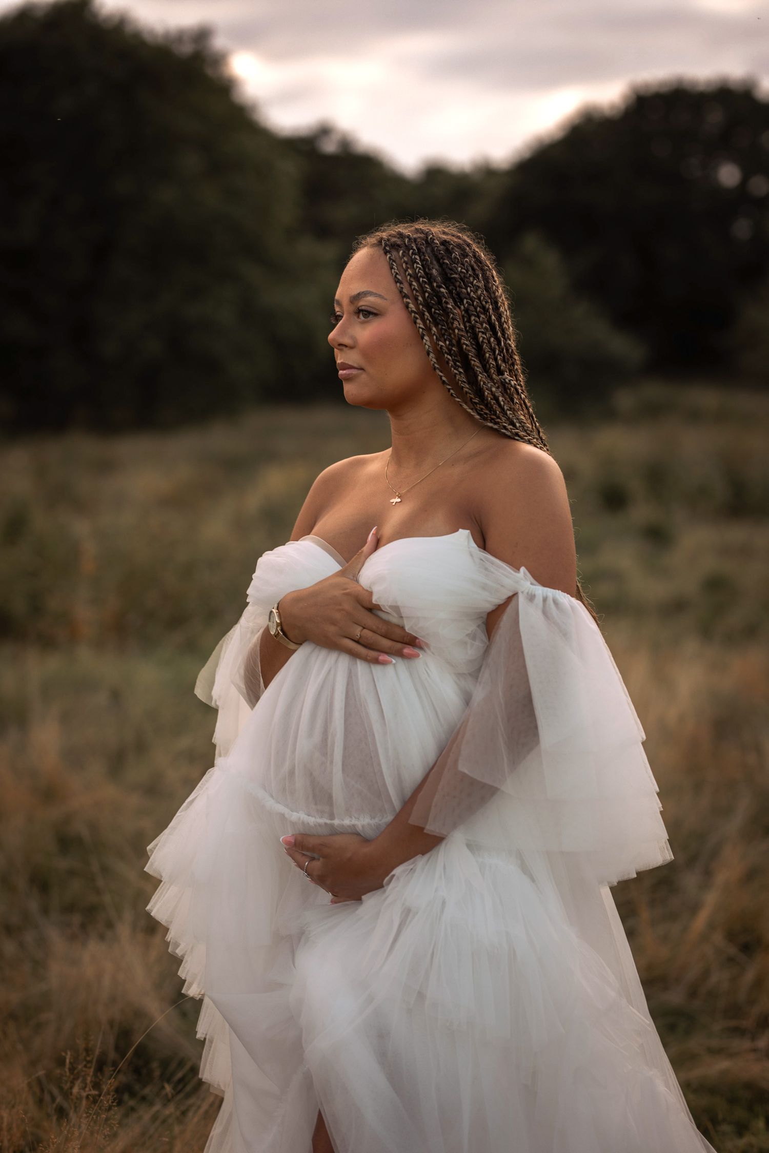 gravid kvinna står på en äng iklädd en vit tyllklänning. hon håller om sin gravidmage.