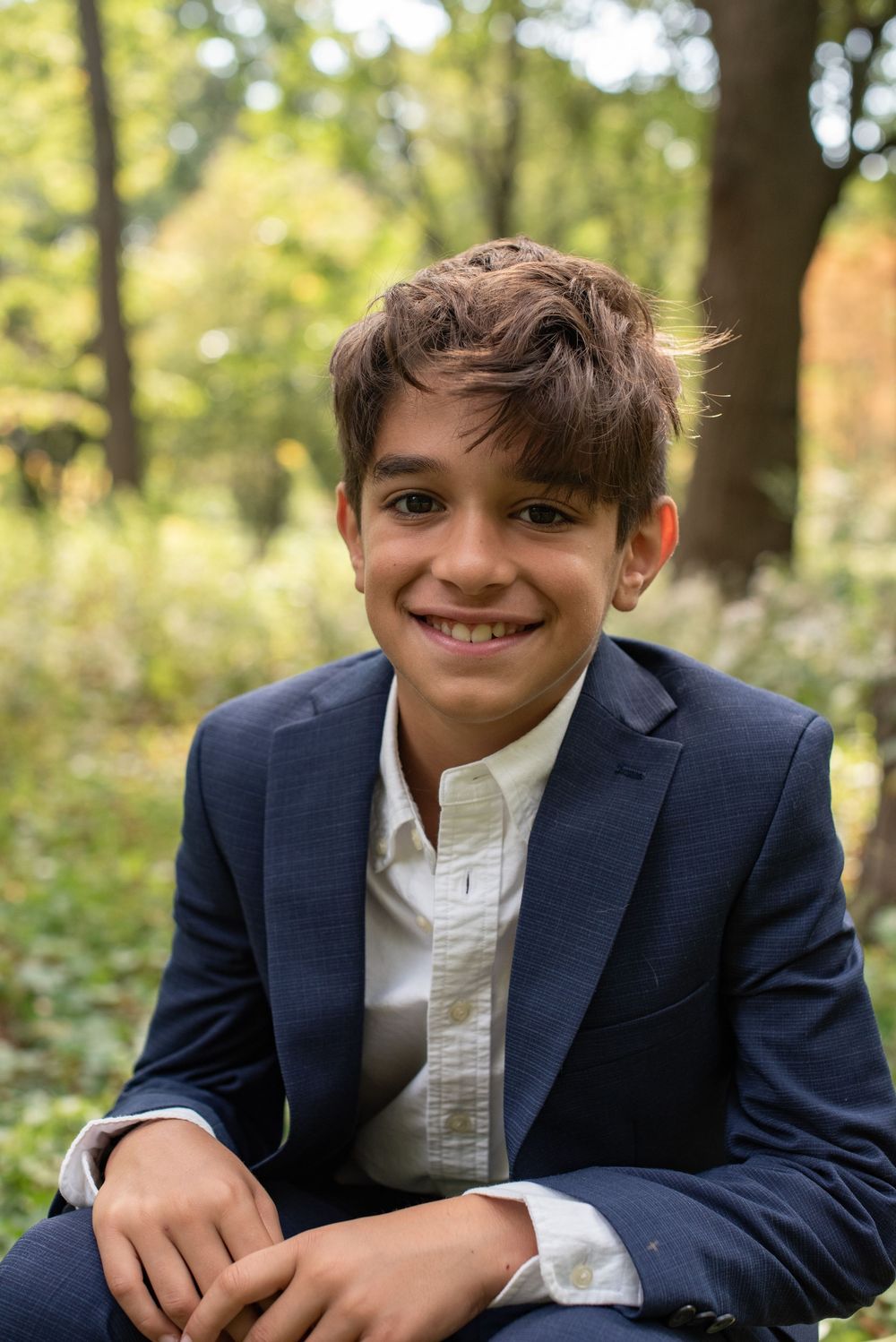 A young person in a navy blue blazer and white shirt poses outdoors against a natural autumn background.