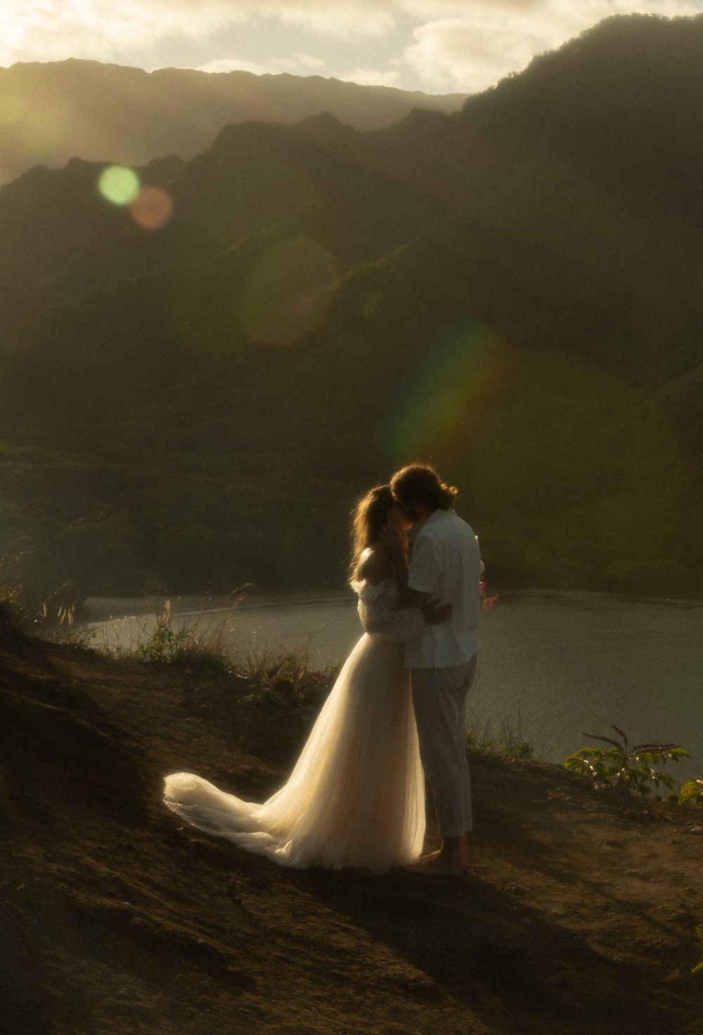 Couple embraces on a mountain overlook at sunset with flowing white wedding dress and lens flare in the background.