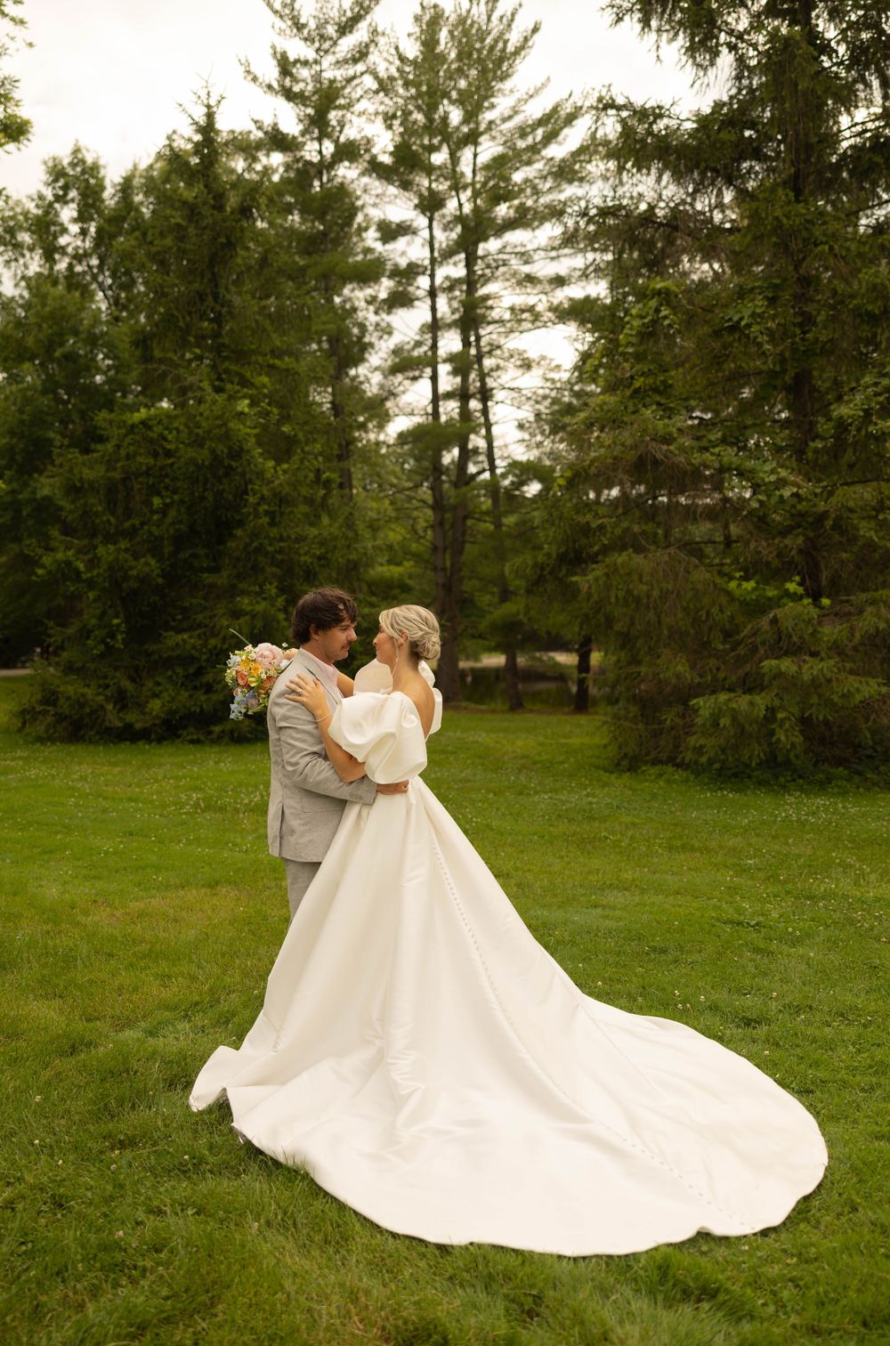 Bride in flowing white dress and groom in gray suit embrace on a lush green lawn.