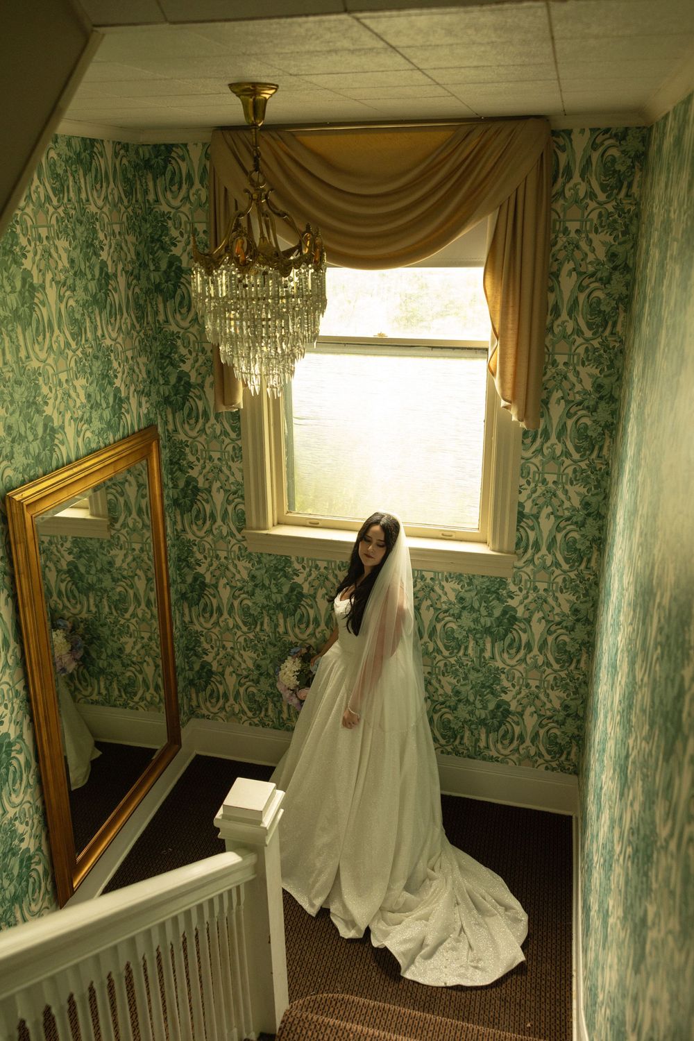 A bride in a white wedding gown and veil poses on vintage staircase with green damask wallpaper and ornate gold mirror.
