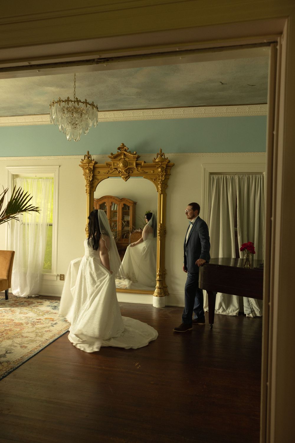 Two figures in an elegant room with ornate gold mirror and chandelier stand on wood floors before the reflection.