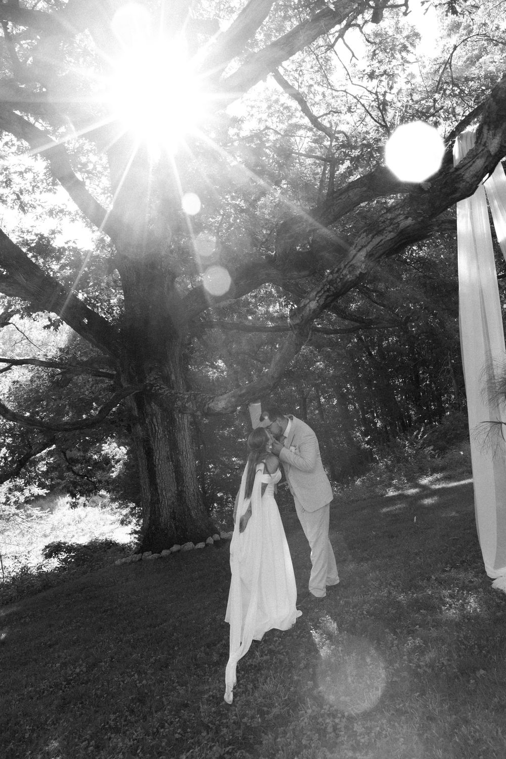 A black and white outdoor wedding photo with sunlight streaming through trees creating a dreamy lens flare effect.