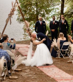 An outdoor wedding ceremony under a rustic wooden arch by a lake with guests watching.