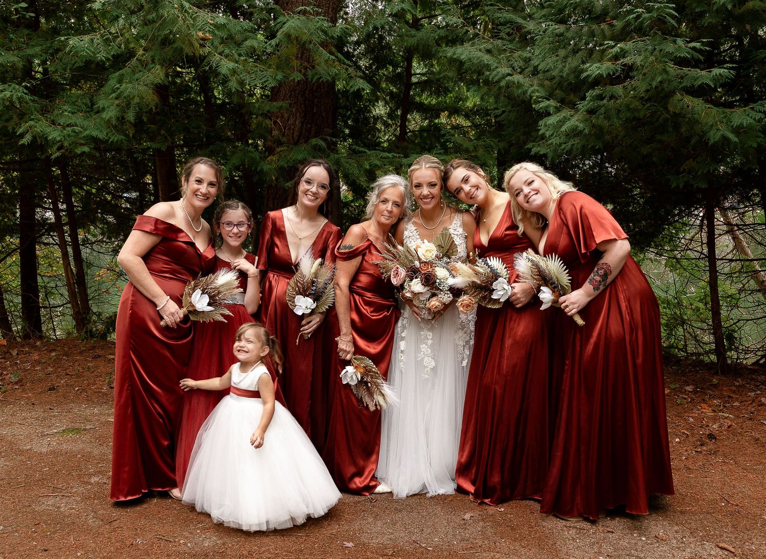 Bridesmaids in burgundy velvet dresses pose with the bride in white against a forest backdrop.