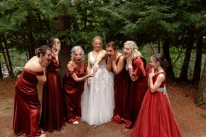 Wedding party in deep red velvet dresses gather around bride for fun group photo in the woods.