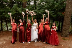 Wedding party in red velvet dresses pose with bouquets raised in a wooded outdoor setting.