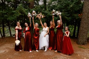 Bridal party in burgundy dresses celebrating with bouquets raised high in a forest setting.