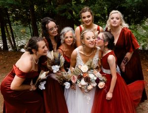 Joyful bridesmaids in red velvet dresses gather close for candid photo in forest setting.