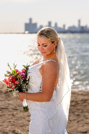 A bride in white lace holds a vibrant bouquet on a beach with city skyline in background.
