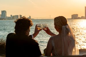 Couple toasts with wine glasses against sunset waterfront view.