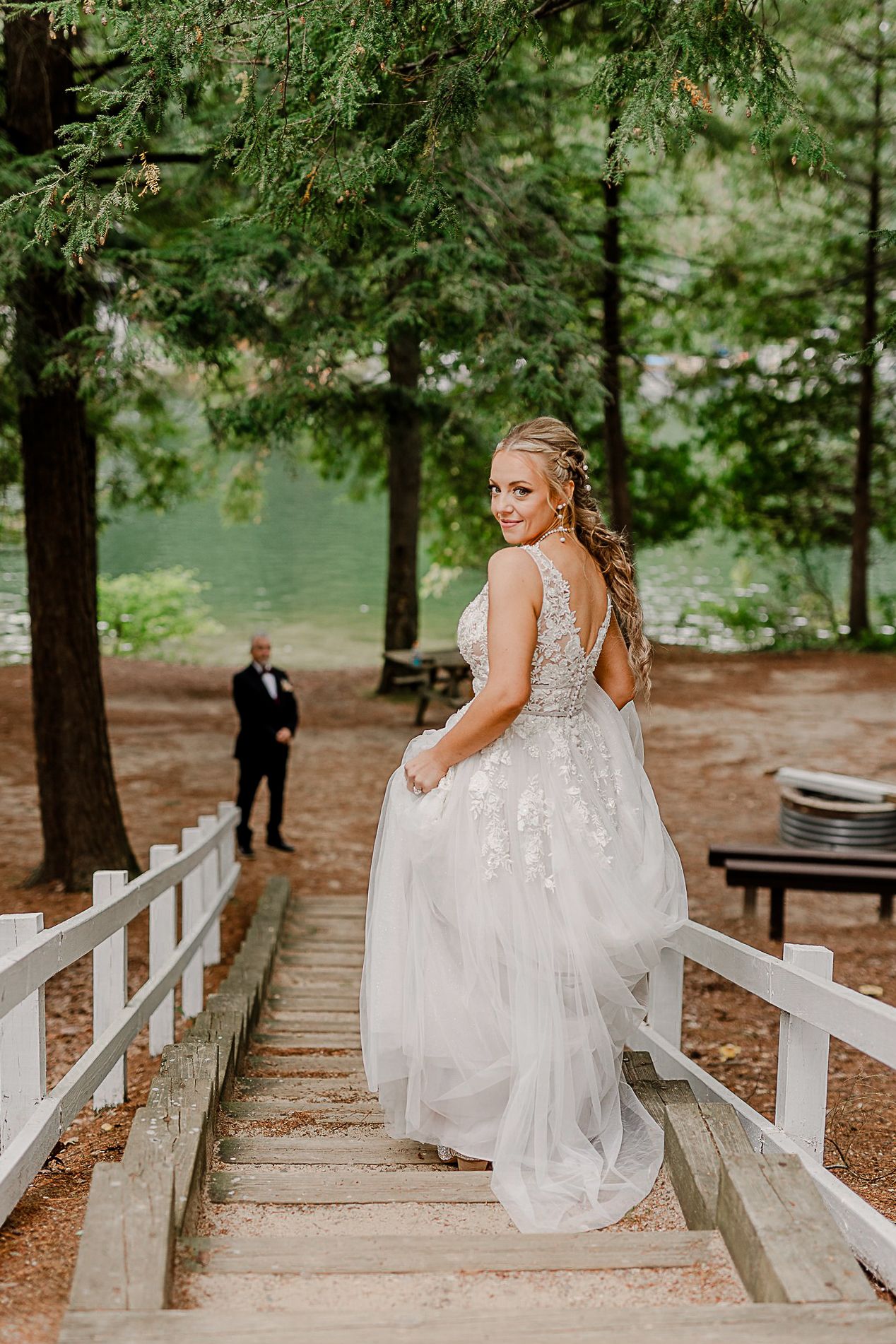 A bride in a flowing lace dress poses on wooden steps surrounded by pine trees.