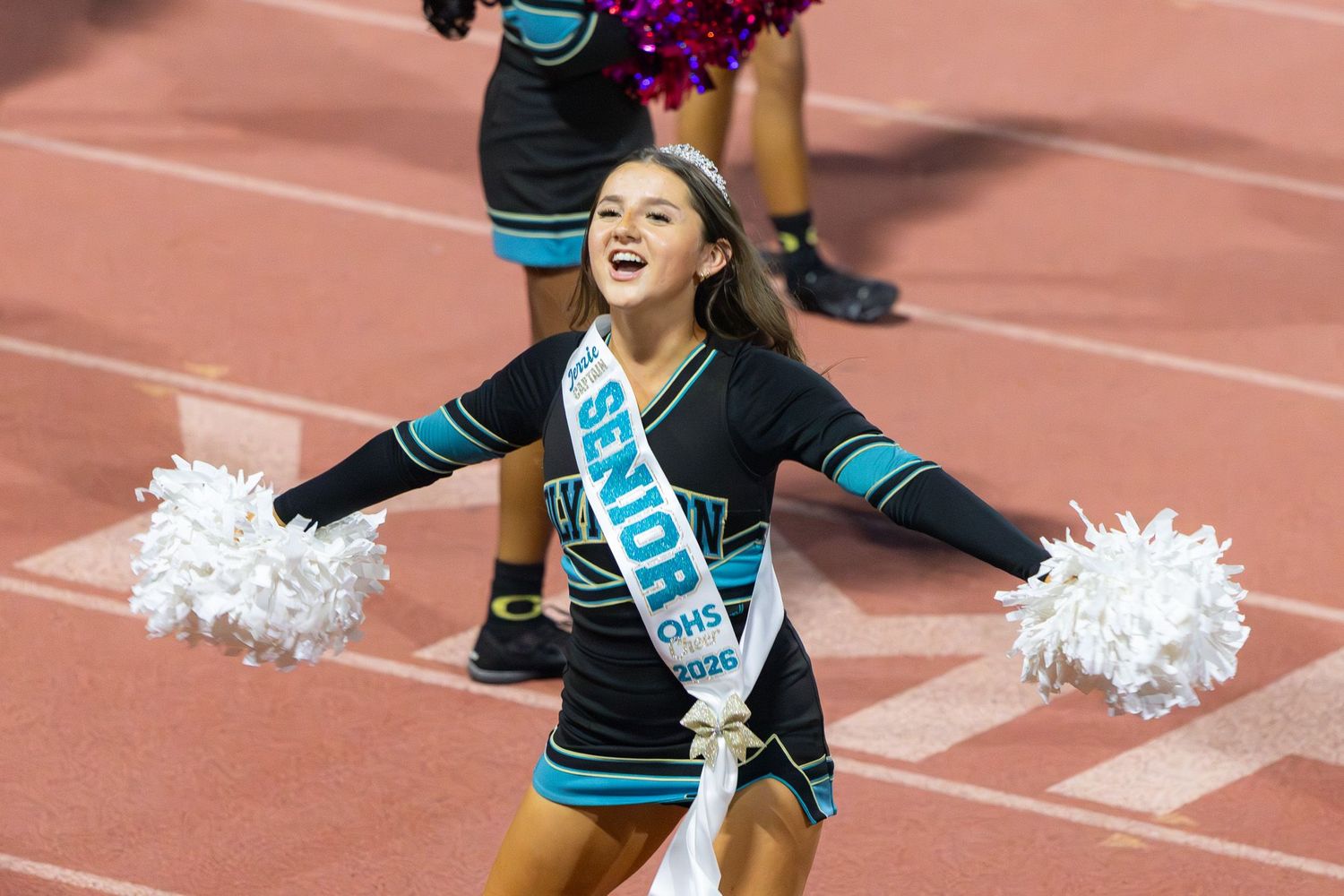 San Diego sports photography of a high school cheerleader wearing a senior sash and holding white pom-poms on a running track.