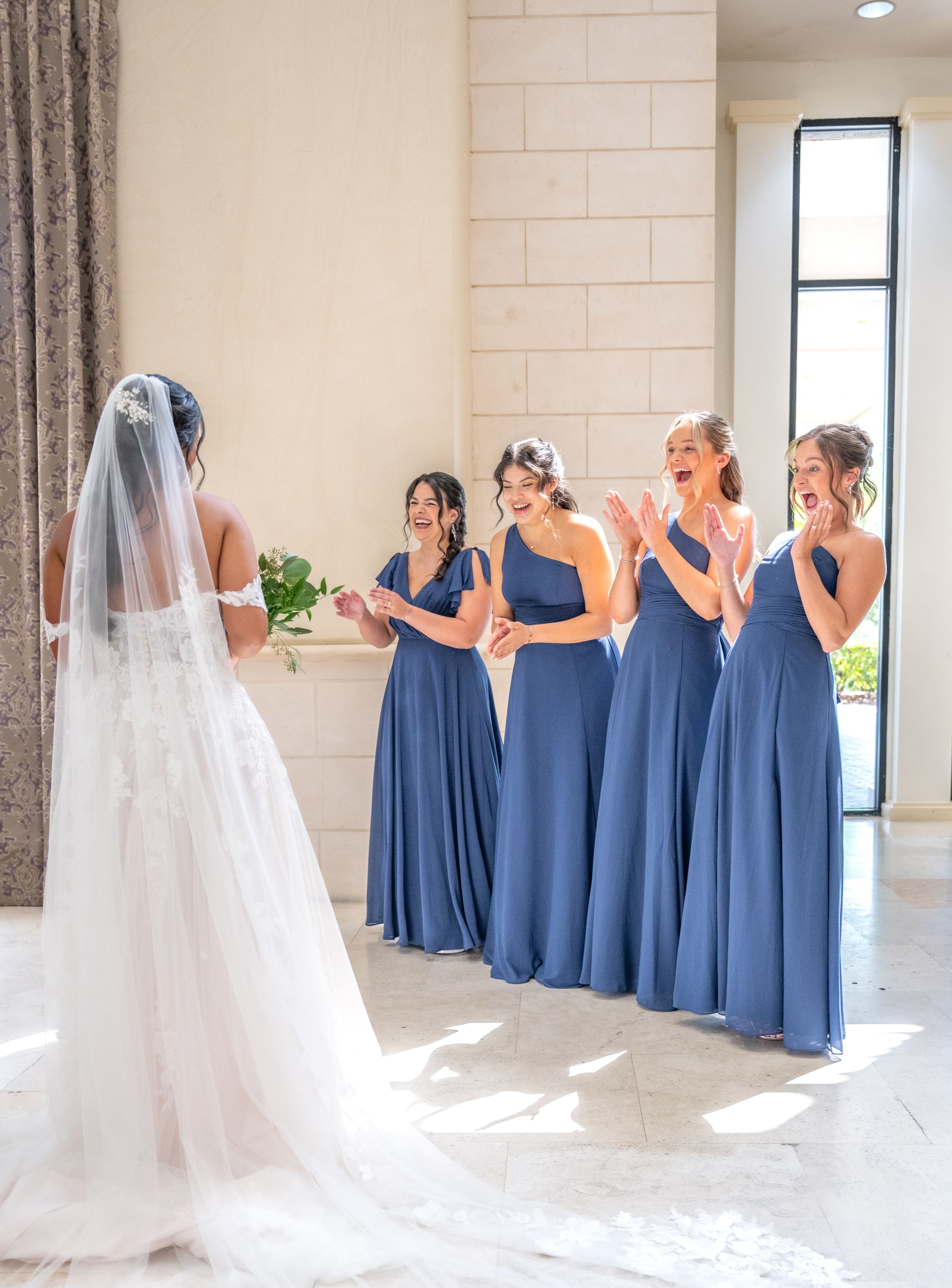Bride in white gown and veil reveals look to four excited bridesmaids in blue dresses during wedding day first look.