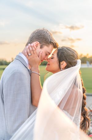 Bride and groom share a romantic forehead touch at sunset, her veil flowing in the breeze on their wedding day.