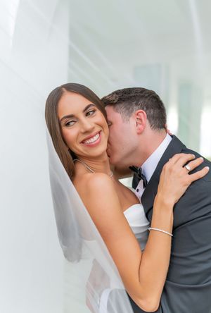 Bride and groom share intimate moment during wedding photoshoot with white veil and formal attire.