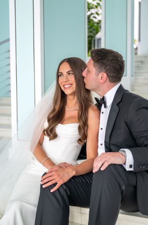 A bride in a white strapless gown and veil sits with a groom in a black tuxedo sharing an intimate moment together.