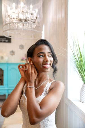 Beautiful bride in white lace dress smiles while putting on jewelry under crystal chandelier.