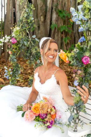 A white lace wedding dress and vibrant floral bouquet create a romantic garden setting with climbing roses and greenery.