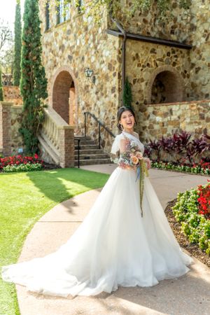 Elegant bride in lace gown poses joyfully at stone venue with blooming garden.