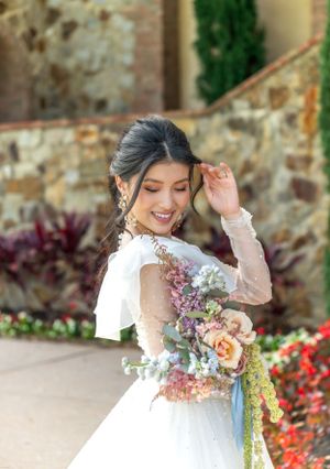 Bride in flowing white dress holds colorful bouquet against stone architecture.