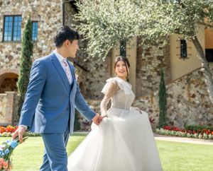 Couple in wedding attire walks through manicured garden with stone building backdrop.