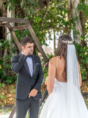 A groom becomes emotional during first look with bride in her backless wedding dress and veil in a garden setting.