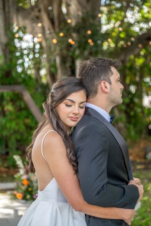A couple in wedding attire share an intimate moment outdoors with string lights and greenery in the background.
