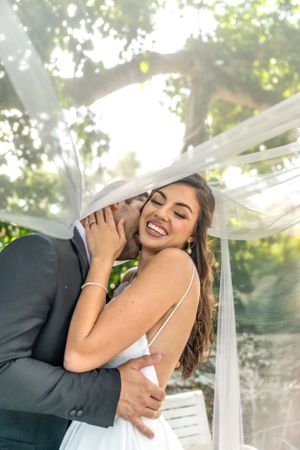 A joyful bride laughs and embraces with her partner during an outdoor wedding photoshoot with flowing veil.