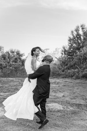 A joyful wedding couple twirls together in black and white on outdoor grass with trees in the background.