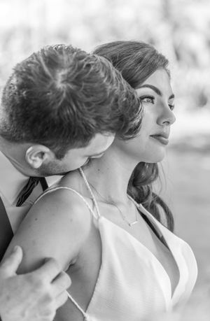 Black and white intimate portrait of a couple in formal attire, with one looking sideways while the other leans in close.