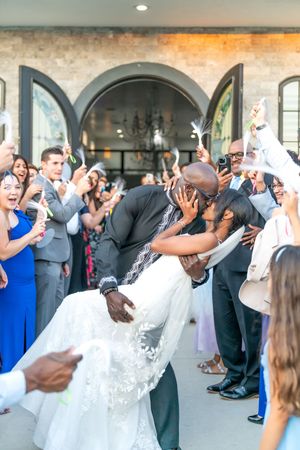 A romantic wedding celebration as guests cheer and applaud while a couple shares a dramatic dip kiss outside an arched venue.