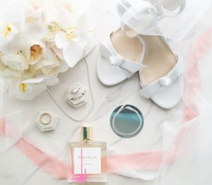 Wedding day accessories flat lay with white shoes, perfume bottle, orchids and jewelry on a soft pink background.