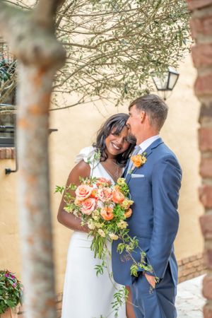 Wedding couple shares intimate moment in courtyard with peach and coral flower bouquet.