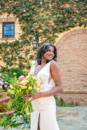 Elegant bride in white dress holds peach and yellow floral bouquet against ivy-covered brick wall.
