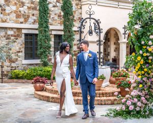 Wedding couple walks together on stone patio with decorative lighting and flowering garden.