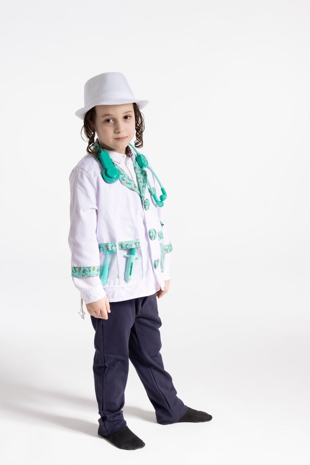 Child dressed as doctor in white coat with teal accessories poses against white background.