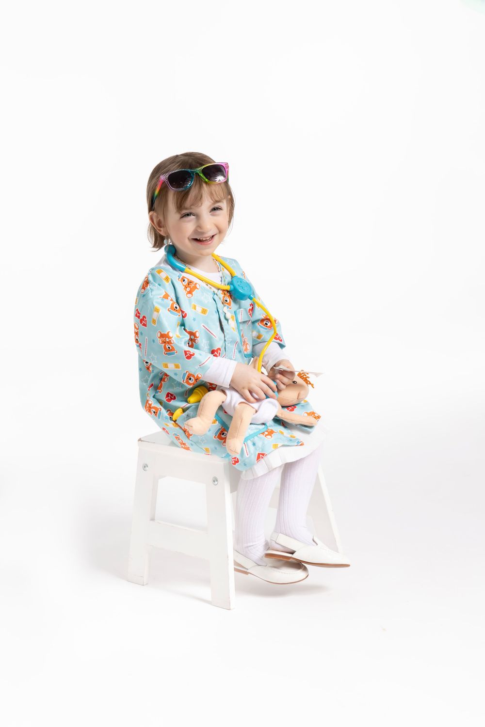 Young child in floral blue medical scrubs sits happily on white stool against white background.