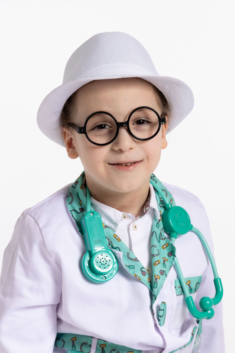 Child wearing round glasses, white hat and medical coat with teal stethoscope smiles at camera.