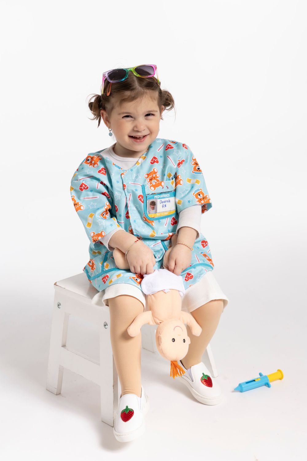 Child wearing blue medical scrubs and sunglasses sits on white stool with toy medical supplies.