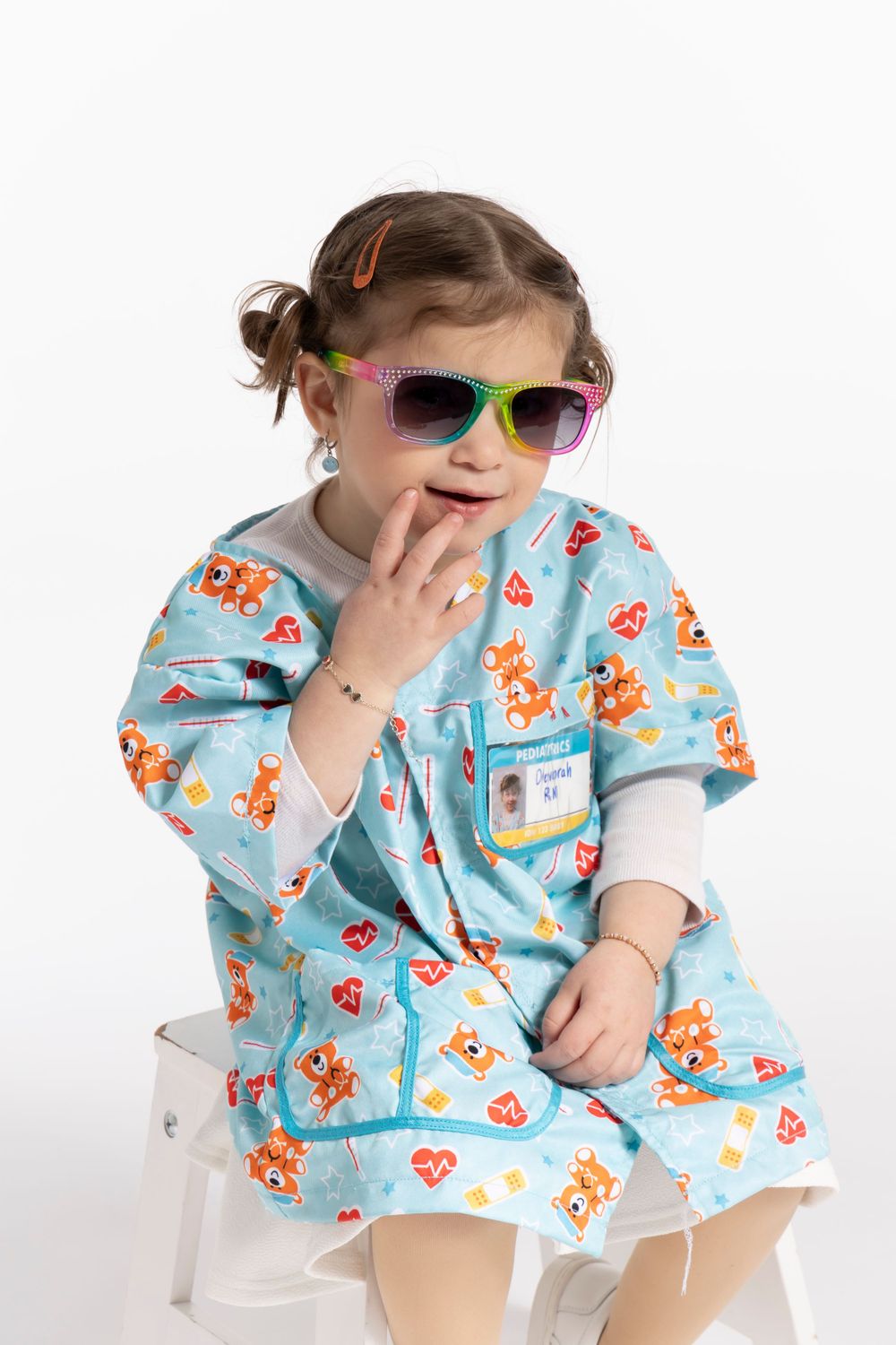 Child in blue medical scrubs and colorful sunglasses poses playfully on white stool.