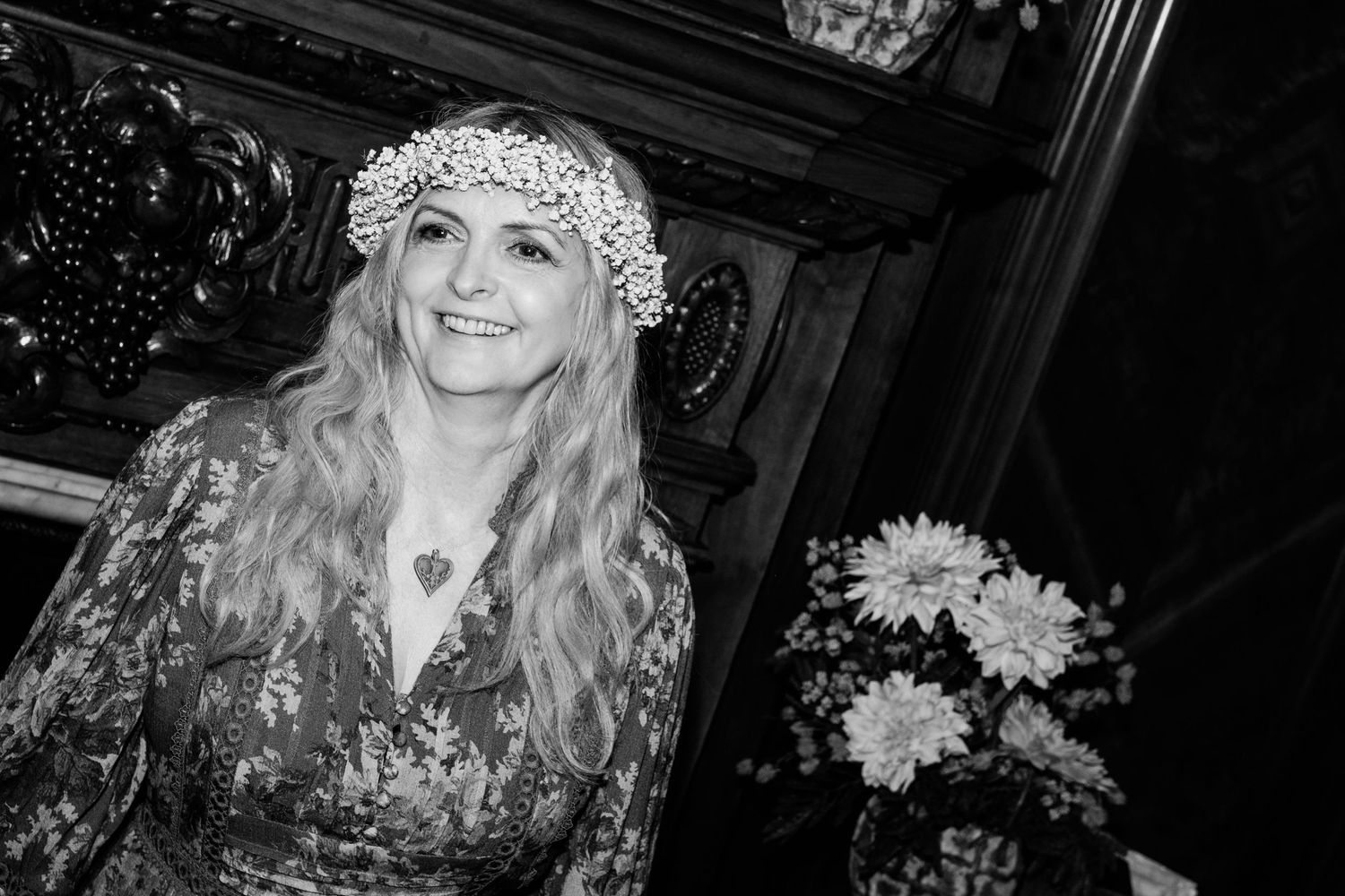 Smiling woman with long blonde hair wearing a floral crown and heart necklace in a black and white photo.