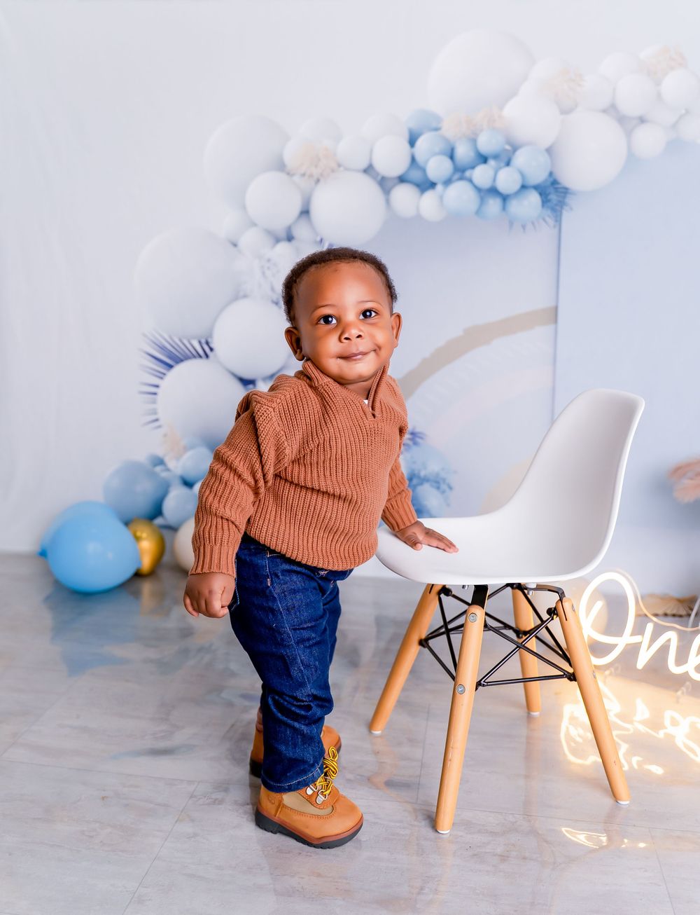 Baby in brown sweater and jeans plays near white chair with blue and white balloon decorations in background.