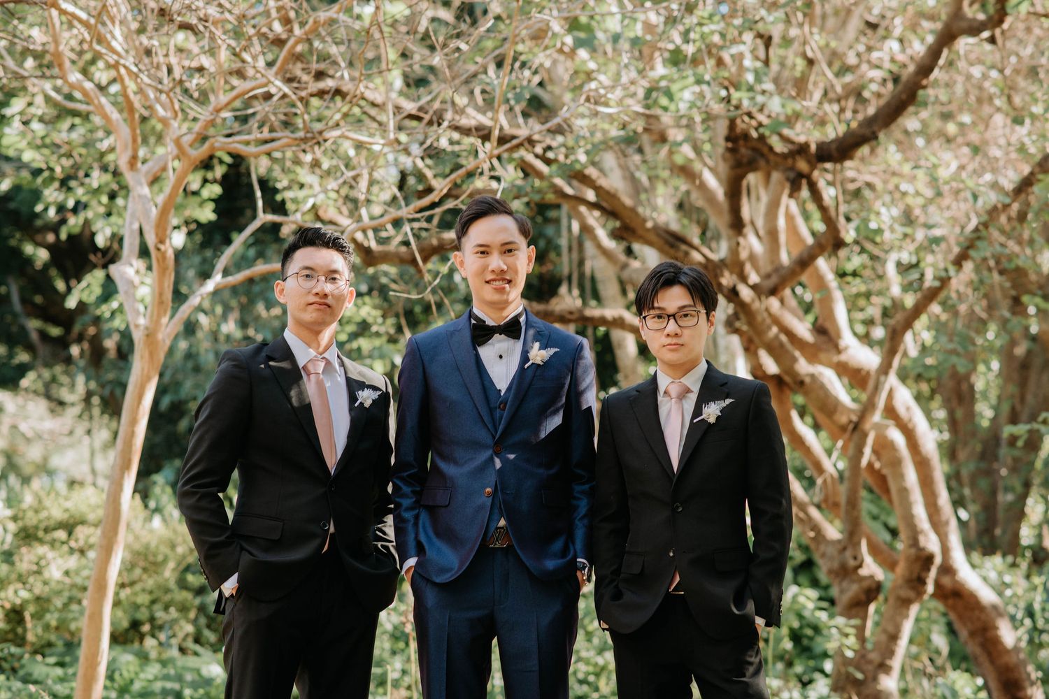 Three men in formal suits pose together in a garden setting.