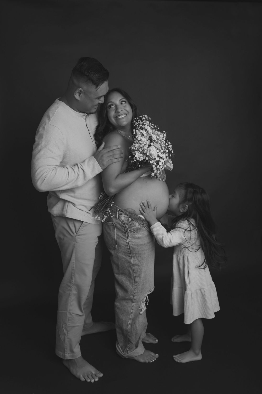 A tender black and white maternity photo showing a couple embracing with a small child touching the pregnant belly.
