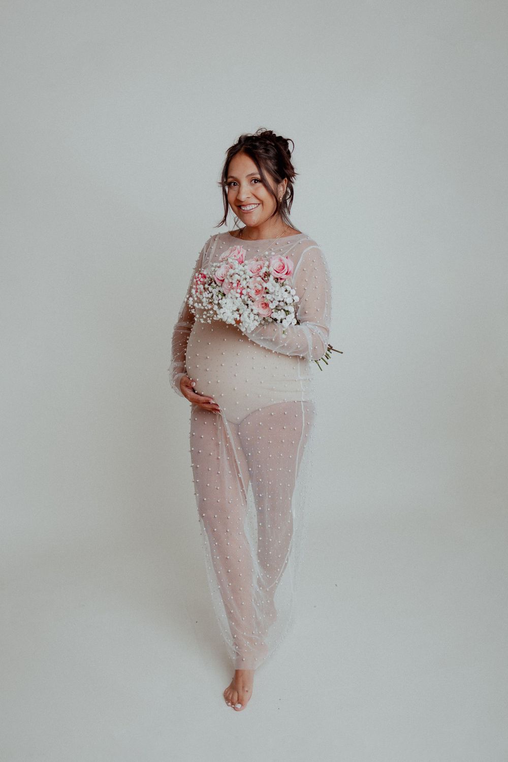 Expectant mother in a sheer lace dress holding delicate pink and white flowers against a light background.