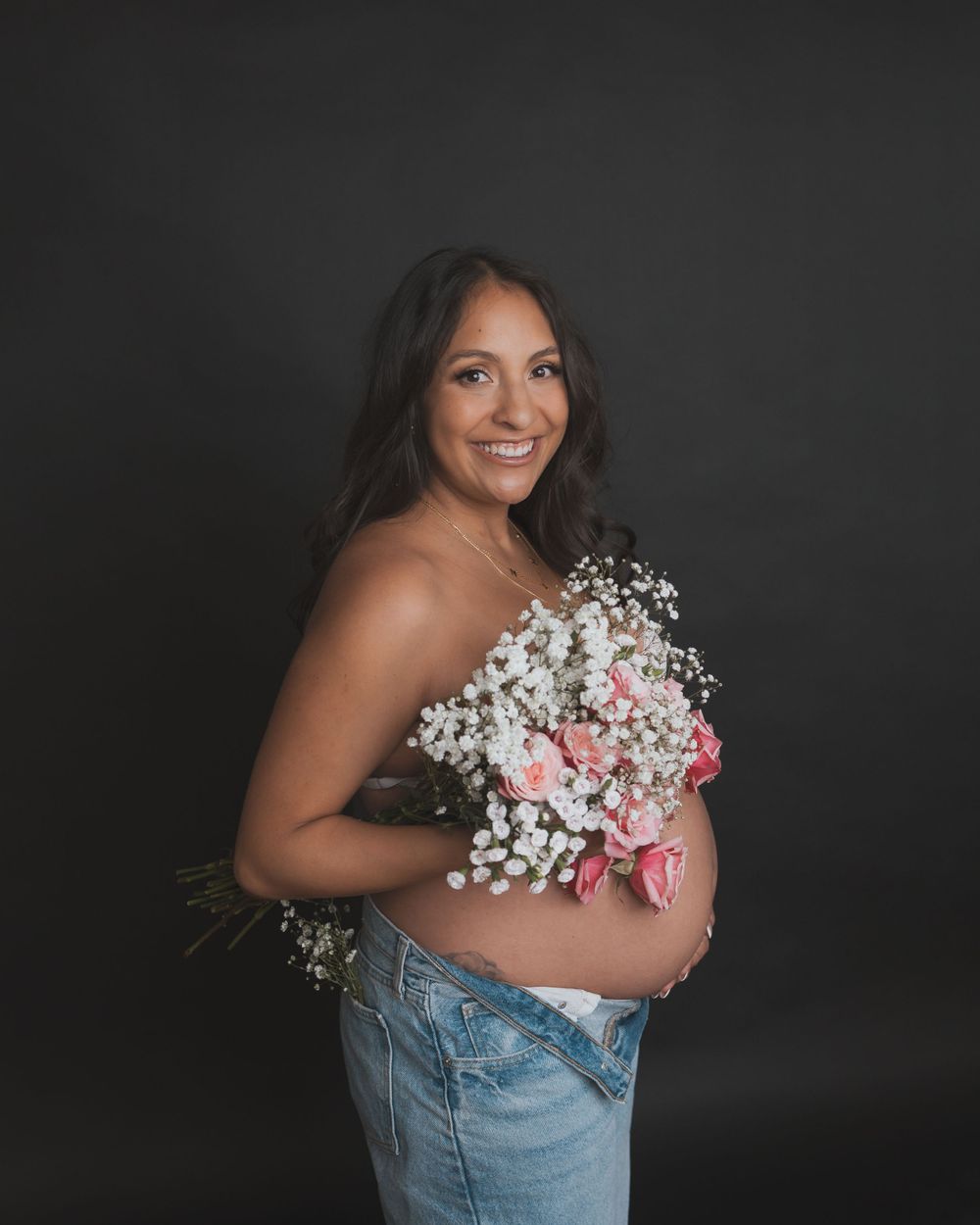 Glowing expectant mother holding a bouquet of baby's breath and pink roses against dark background.